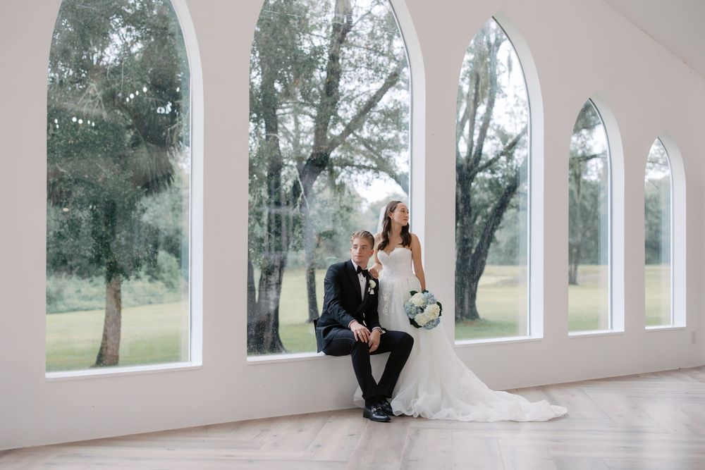 Bride and groom pose elegantly by tall arched windows in a bright white wedding venue with lush green trees outside.
