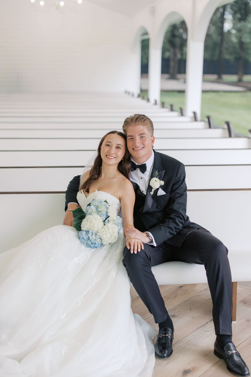 Bride and groom sitting together in white chapel pews, bride holding blue hydrangea bouquet, groom in black tuxedo with bow tie.