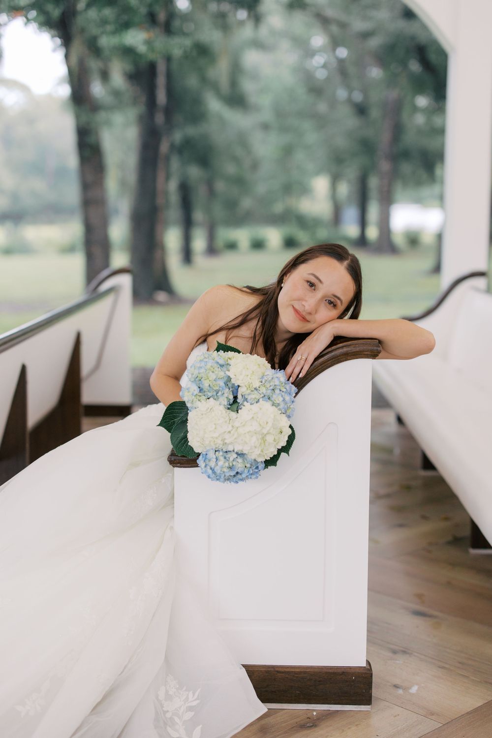 Bride in white gown sitting in church pew holding blue and white hydrangea bouquet, with lush green trees visible through windows.