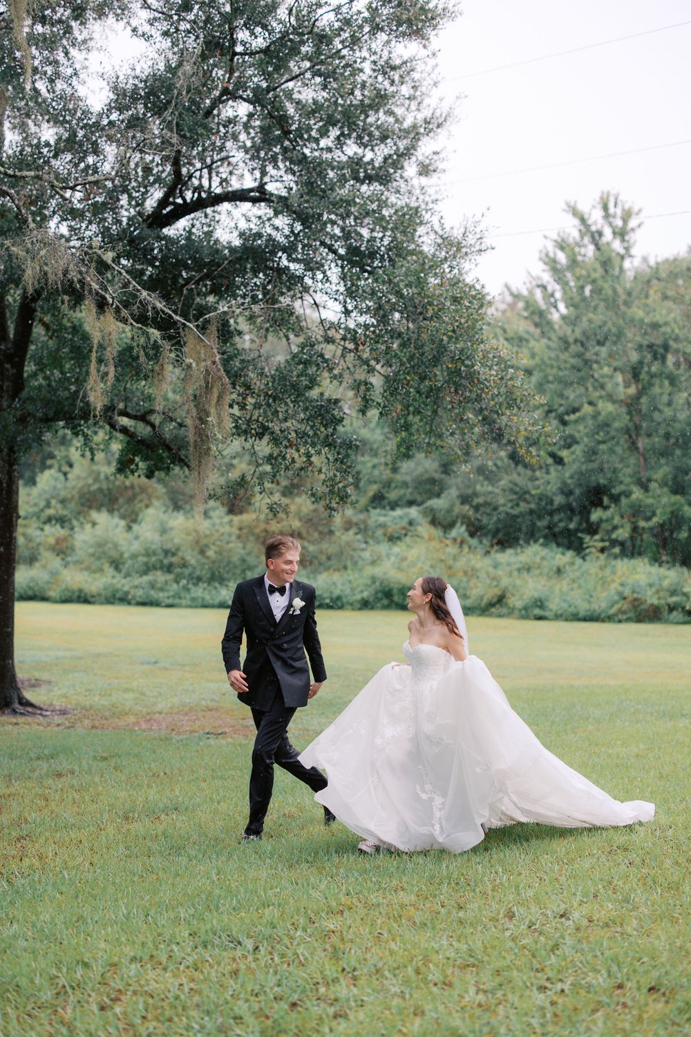 Bride in white ball gown and groom in black tuxedo walk together on lush green lawn surrounded by trees.