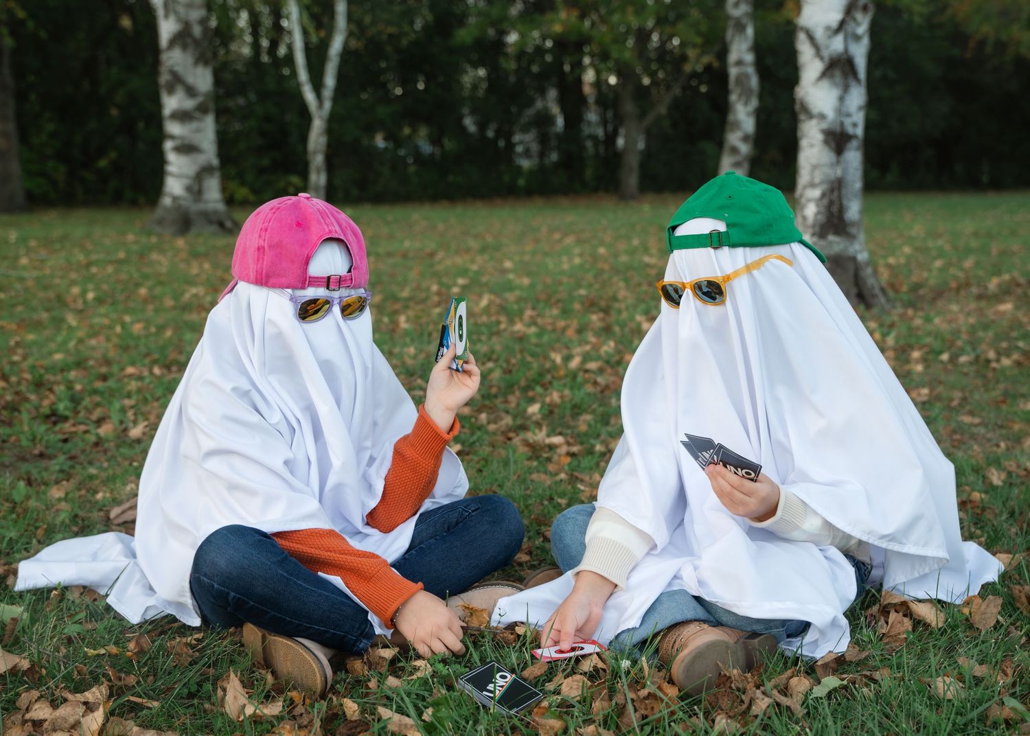 Two people in white sheet ghost costumes, wearing sunglasses and caps, sit on autumn grass looking at their phones.