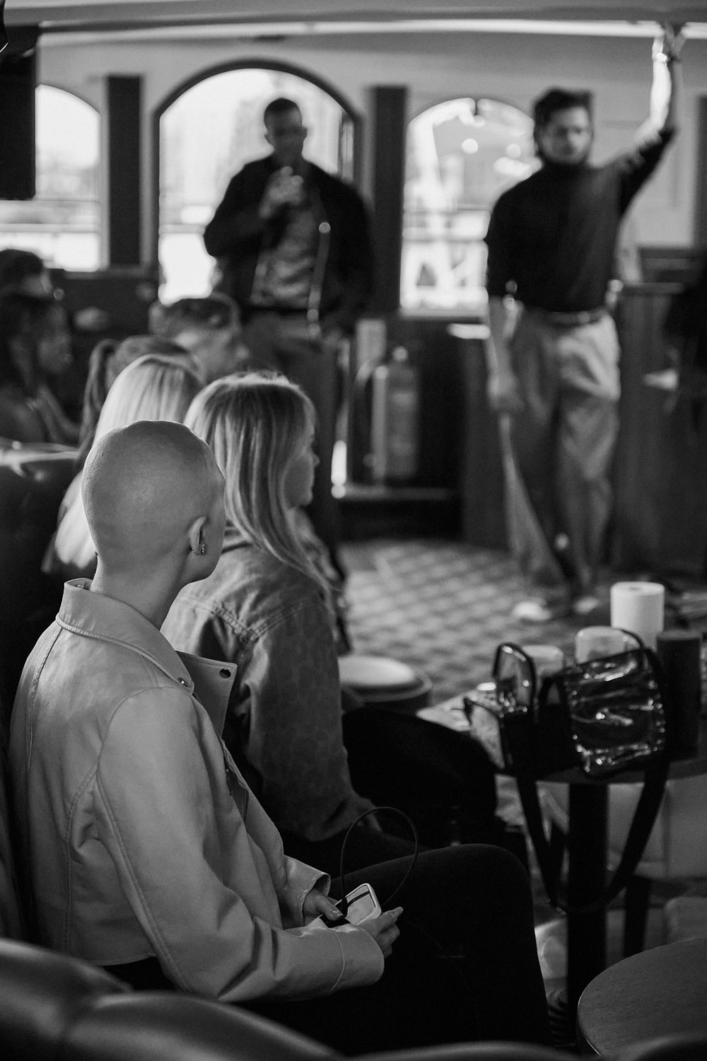 Black and white photo of seated audience members watching two standing men in a room with arched windows and a patterned carpet.