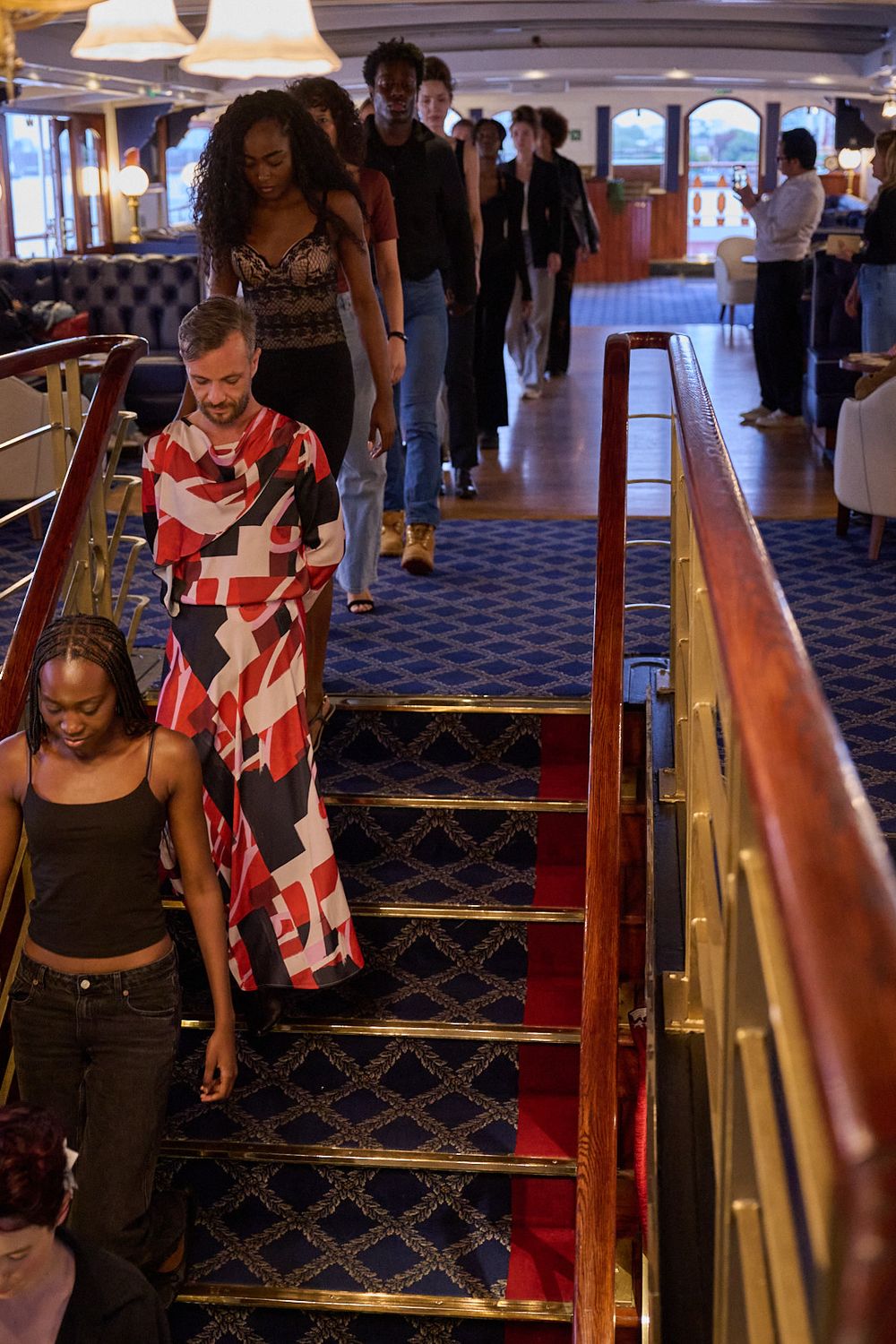 Group of people in stylish outfits walking down a carpeted staircase with brass railings inside an elegant ship-style interior.