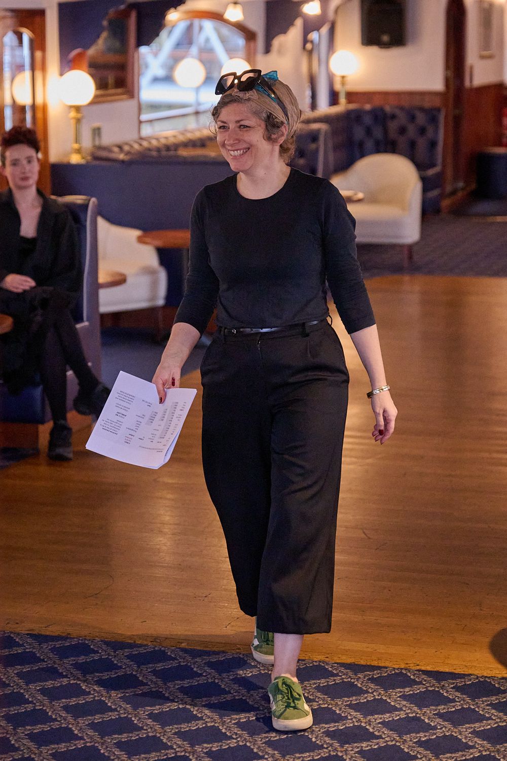 Smiling woman in black top, wide-leg trousers and green sneakers walks across a wooden floor holding papers, with a lounge behind her.