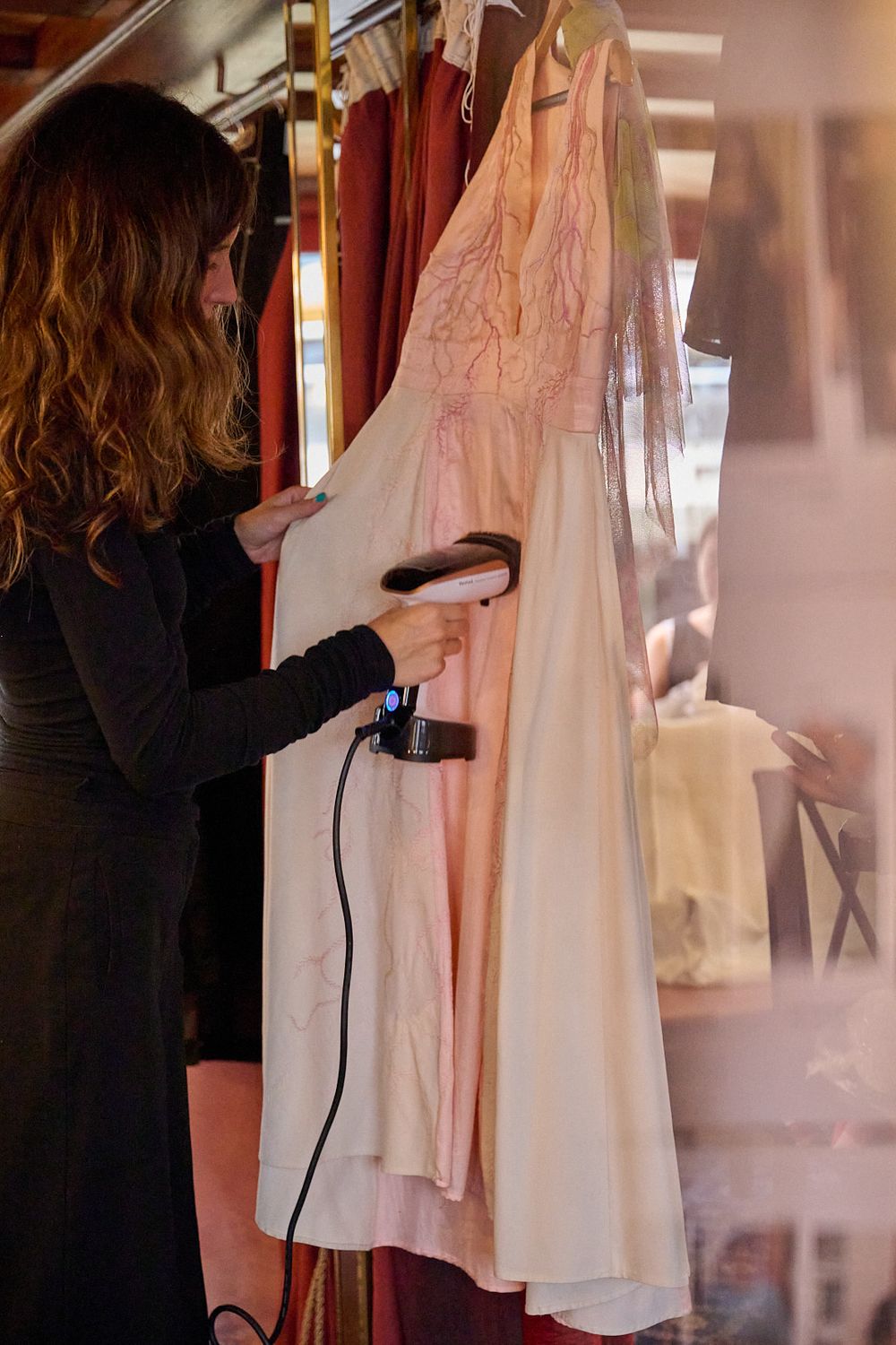Woman with wavy brown hair steaming a delicate cream and blush pink embroidered dress hanging on a rack using a handheld garment steamer.