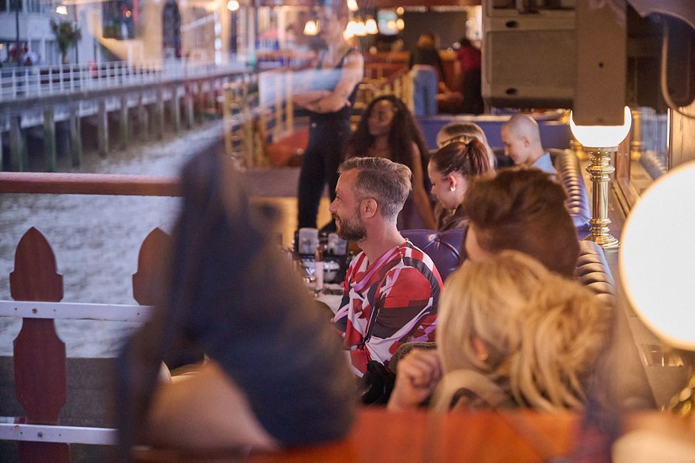 Group of friends laughing and dining together at a lively waterfront restaurant terrace with warm lighting at dusk.