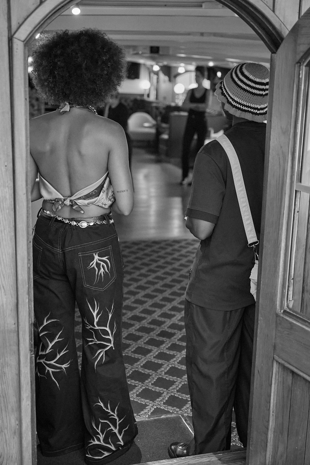 Black and white photo of two people from behind in a wooden doorway, one in a halter top and tree-embroidered jeans, the other in a striped 