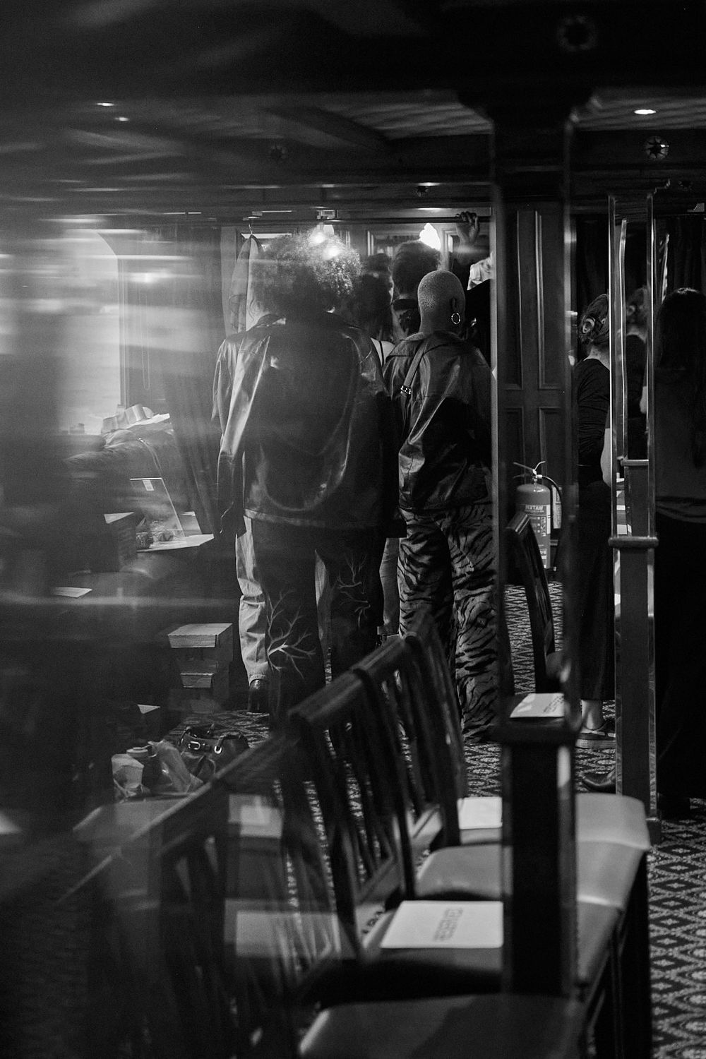 Black and white photo of people standing near rows of chairs and tables in a dimly lit room, with glass reflections adding depth.