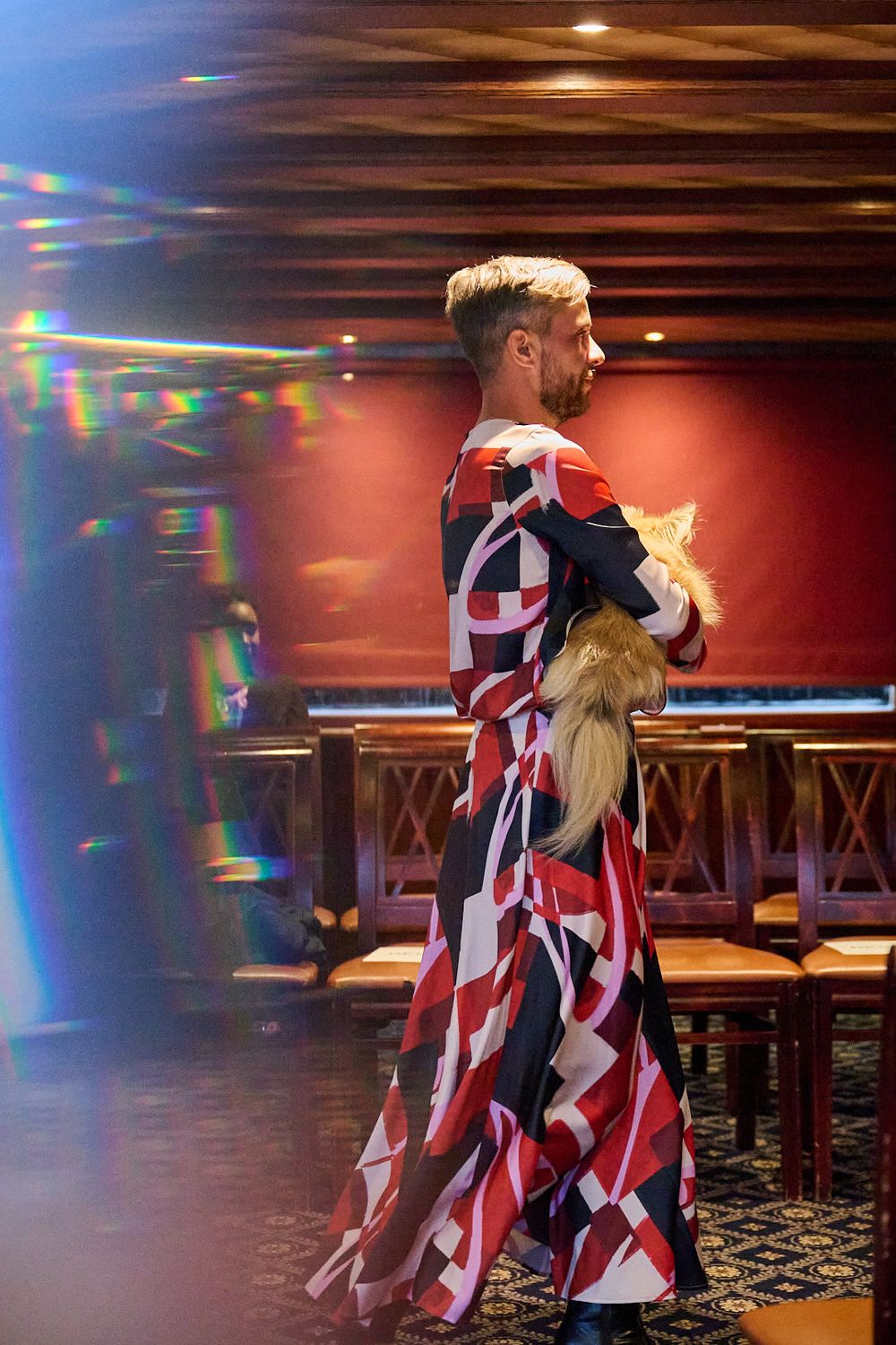 Person in a bold red, white and black geometric maxi dress carrying a fluffy cat through a warmly lit restaurant with wooden chairs.