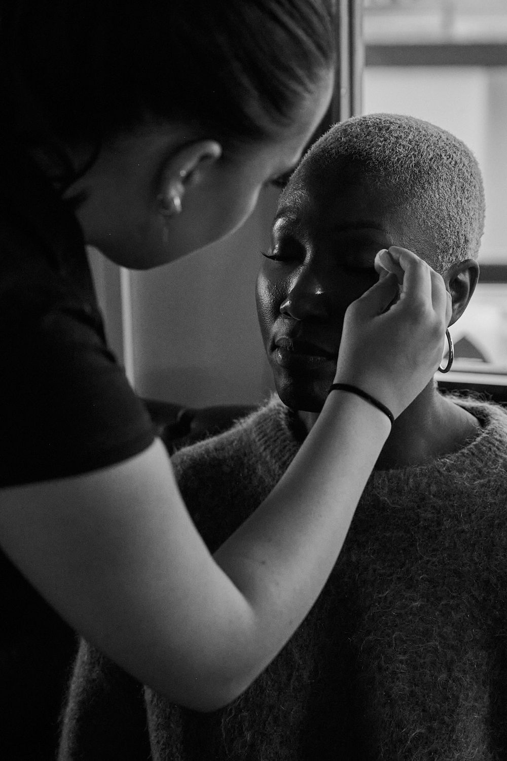Black and white photo of a makeup artist gently applying makeup near the eye of a client with short cropped hair wearing a knit sweater.