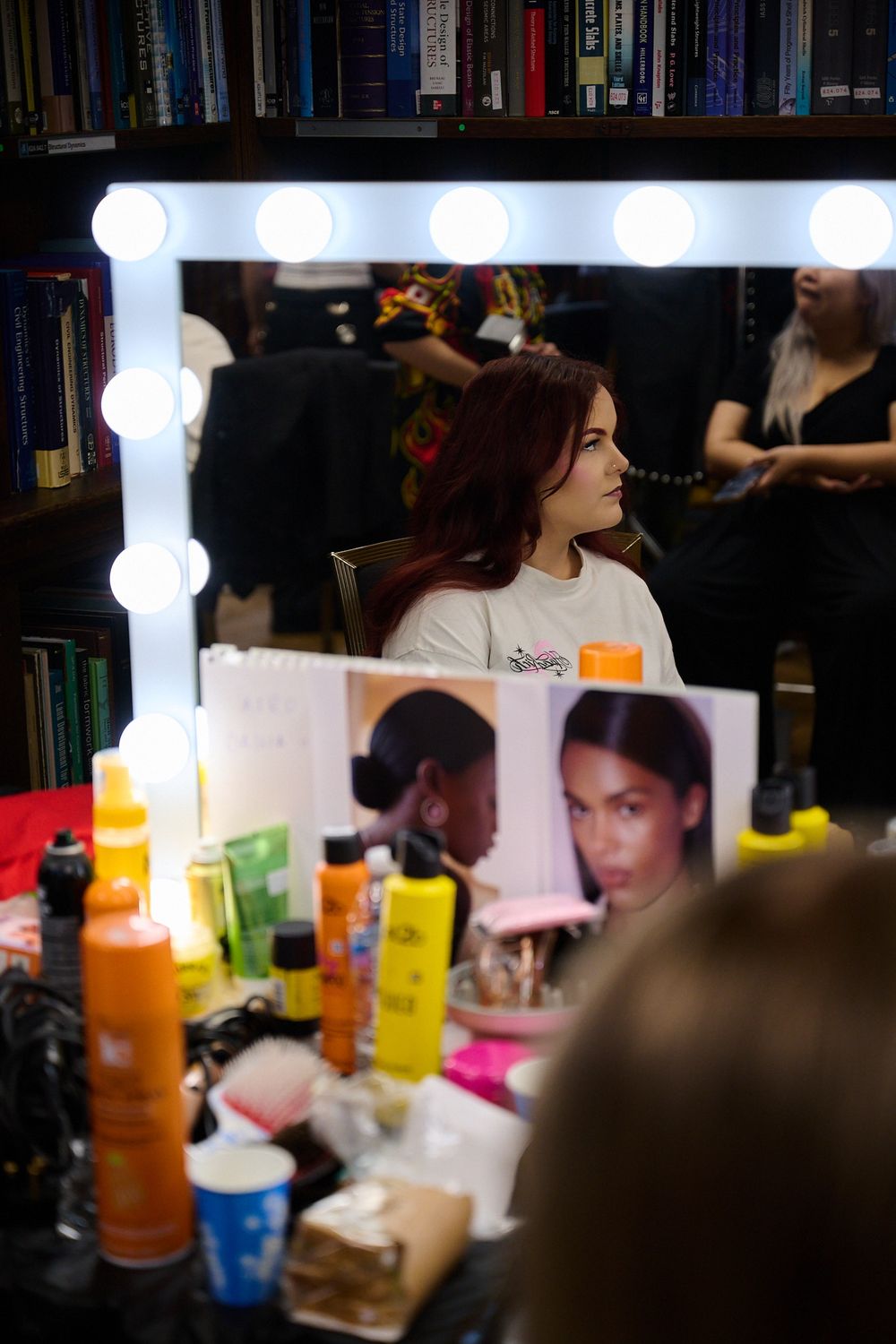 Red-haired woman sits in a hair salon chair, seen in a lit vanity mirror with styling products and beauty magazine photos on the counter.