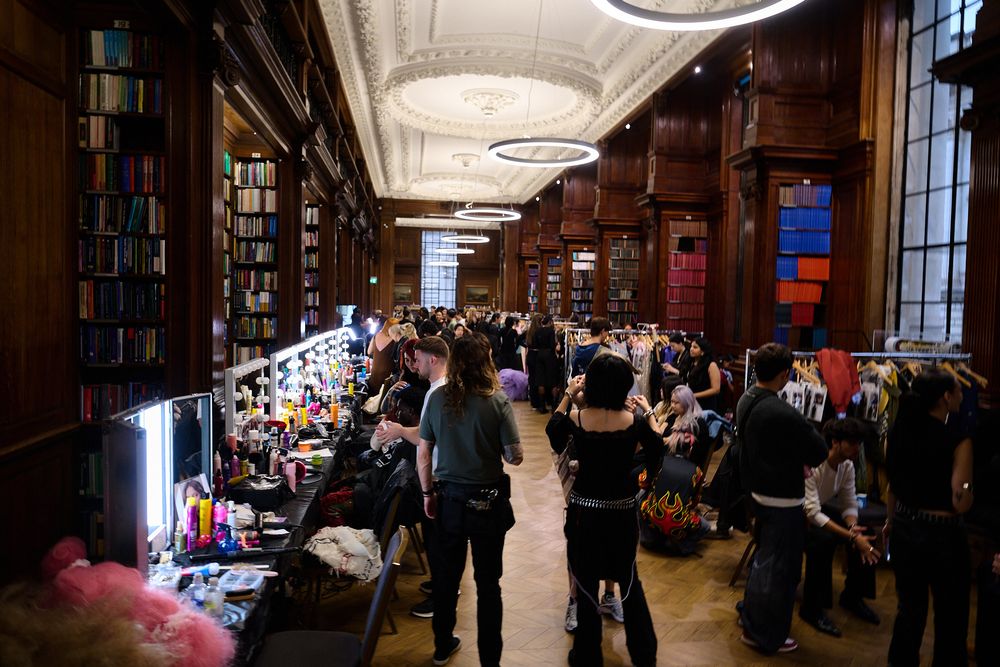 Crowd of visitors browsing vendor tables at a lively event inside a grand wood-paneled library with tall bookshelves and ornate ceilings.