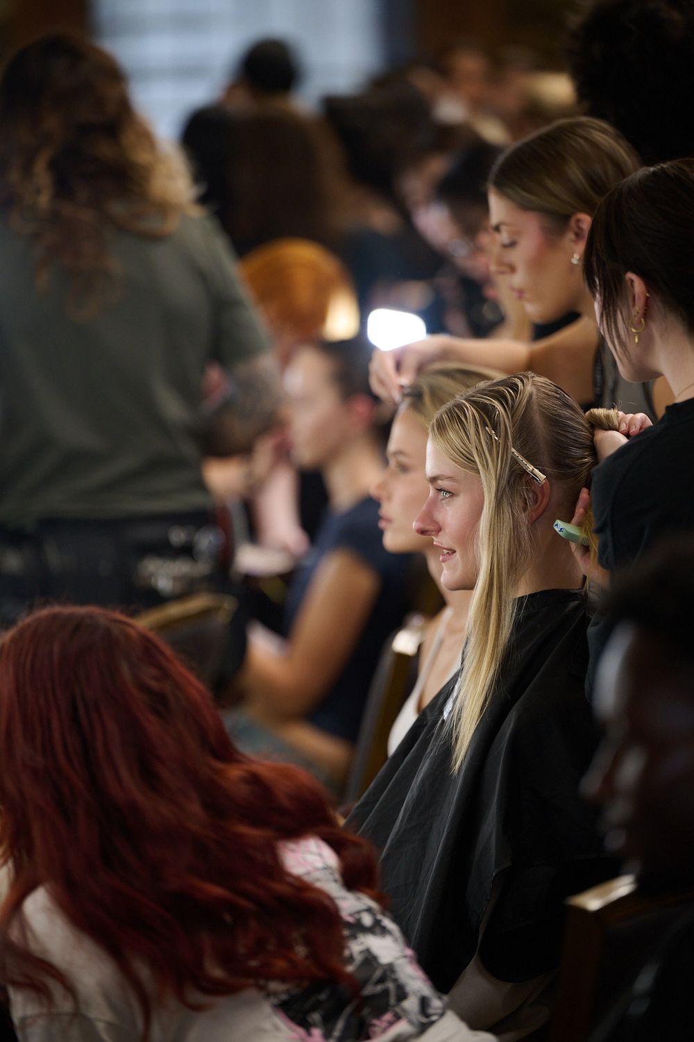 Hairstylists work on models wearing black salon capes in a busy backstage setting, styling blonde and brown hair ahead of a fashion show.