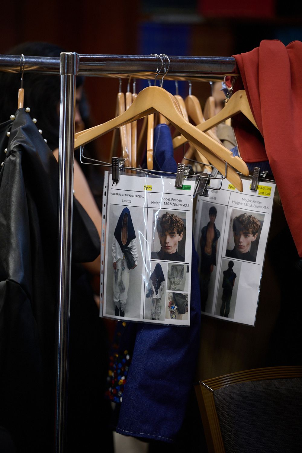 Backstage fashion show clothing rack with wooden hangers, a black leather jacket, and plastic look cards showing model photos.