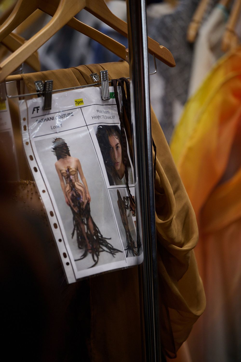 Backstage fashion look card with model photos clipped to a garment rack beside hangers and flowing golden fabric.