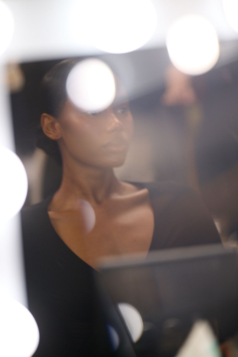 Woman with hair pulled back in a black top seen in a backstage mirror framed by glowing round vanity bulbs.