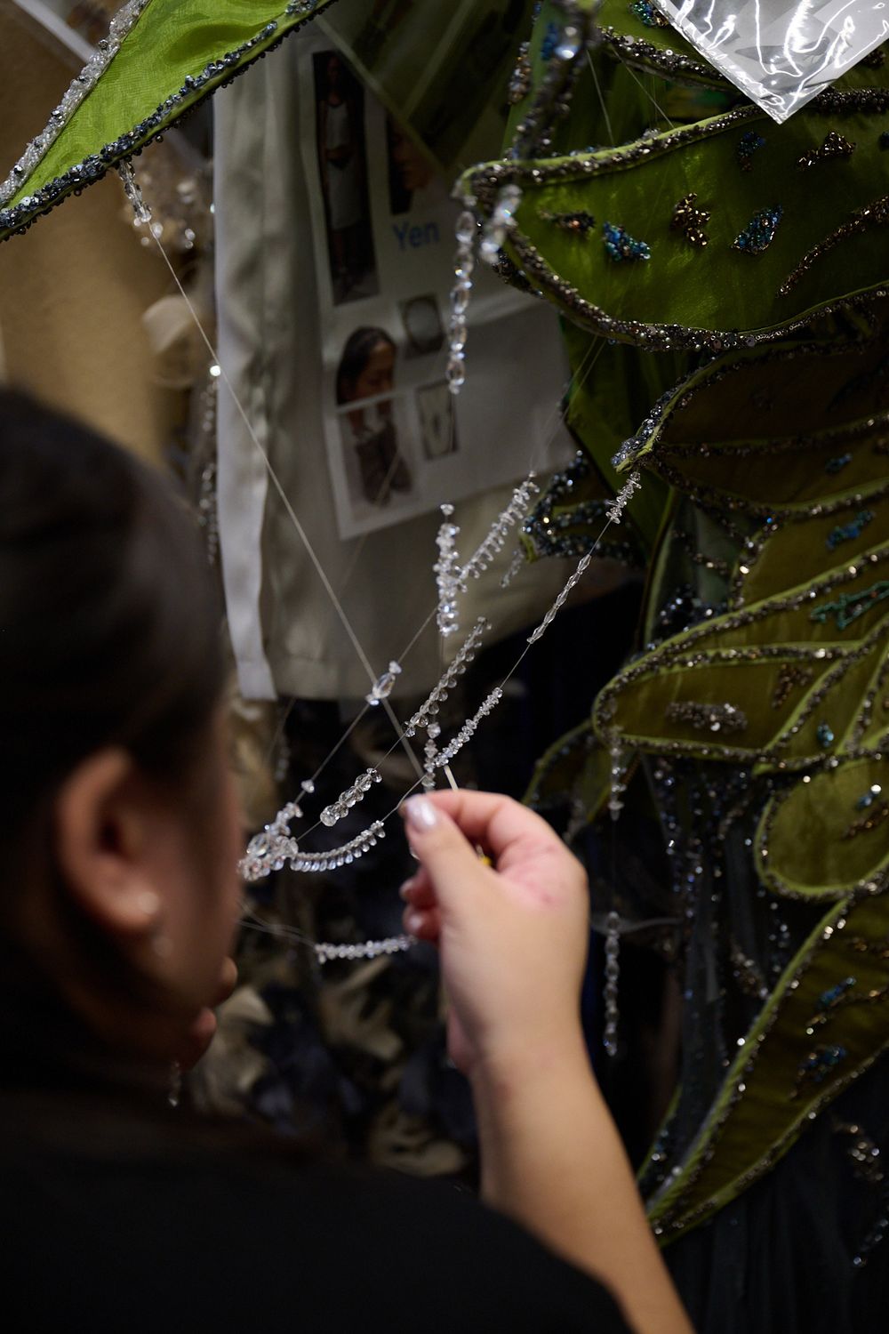 Woman carefully attaches strands of crystal beads to an ornate green costume embellished with sequins and decorative trim.