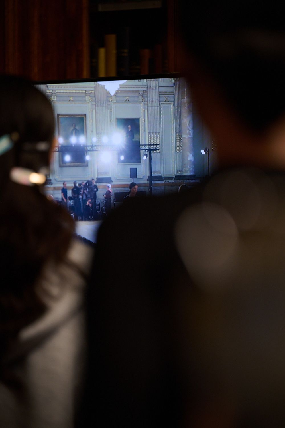 Two blurred silhouettes in the foreground watch a lit screen showing a group of people gathered before an ornate building in a dim room.