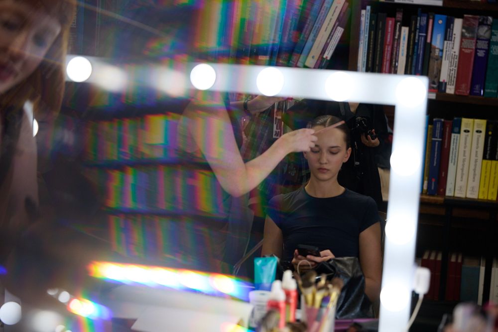 Makeup artist applies cosmetics to a woman seated before a lighted vanity mirror in a studio with bookshelves behind her.