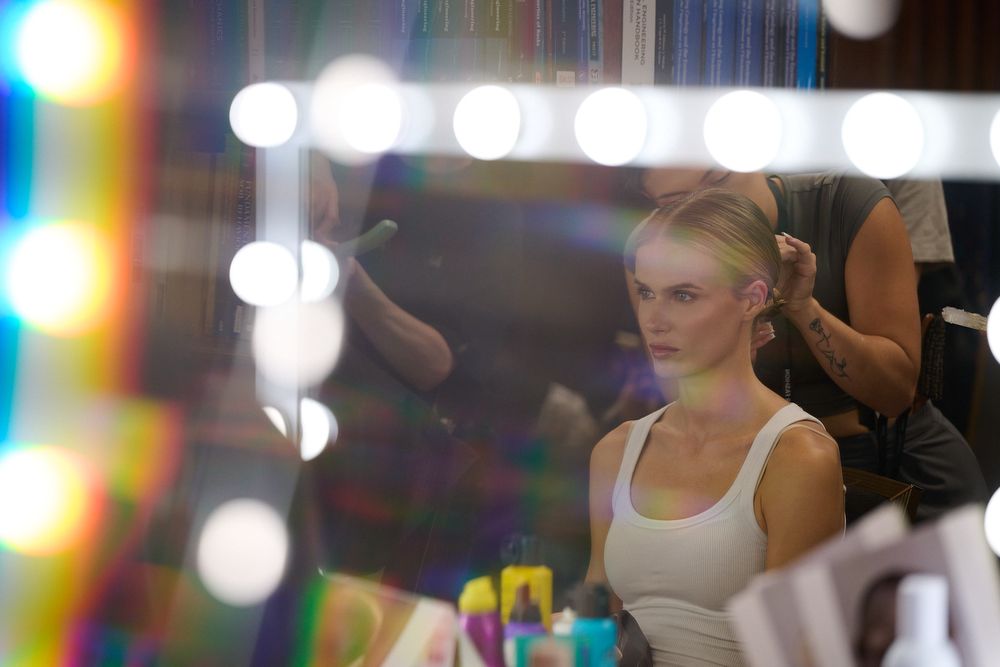 Blonde woman in a white tank top sits before a backstage mirror ringed with bright bulbs while a makeup artist works behind her.