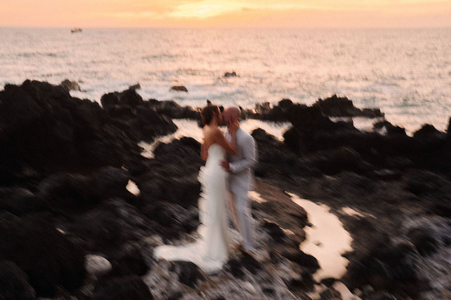 Blurry romantic couple in wedding attire standing on volcanic rocks by the ocean at sunset.