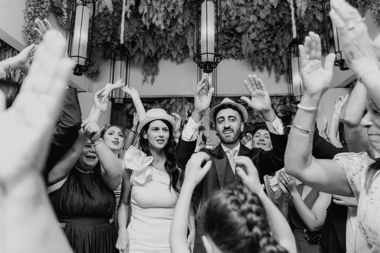 Anteprima della gallery: Lorenzo e Laura con cappelli, tra le mani alzate degli invitati, in bianco e nero.