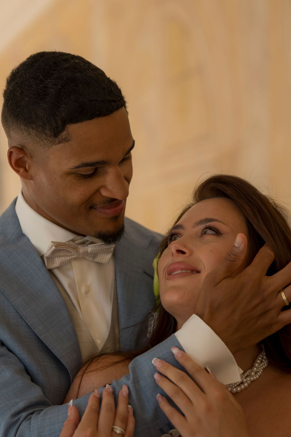 A groom in a blue suit tenderly holds his bride's face as they share an intimate moment at their wedding.