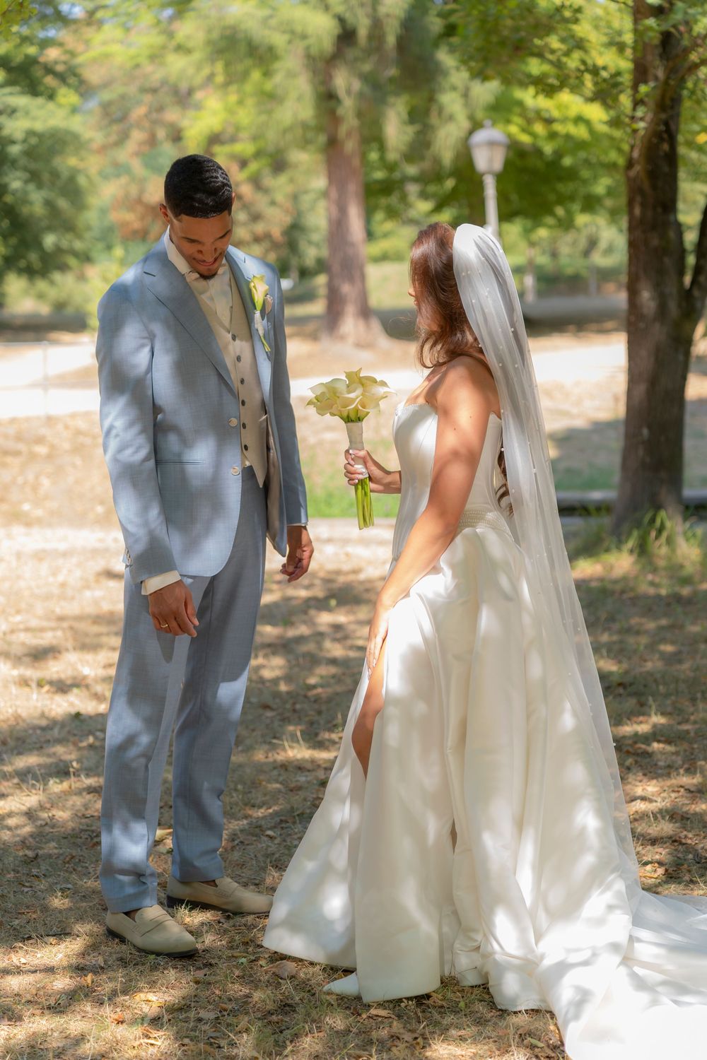 Bride in white gown holding bouquet faces groom in light blue suit during outdoor wedding ceremony in sunny park.