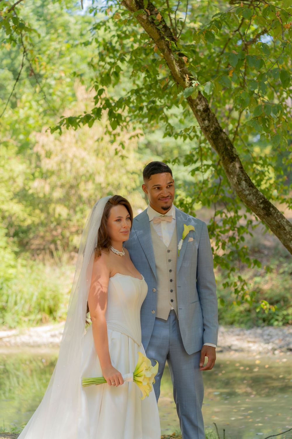 Interracial newlywed couple posing outdoors under green trees, bride in white gown with veil, groom in light blue suit.