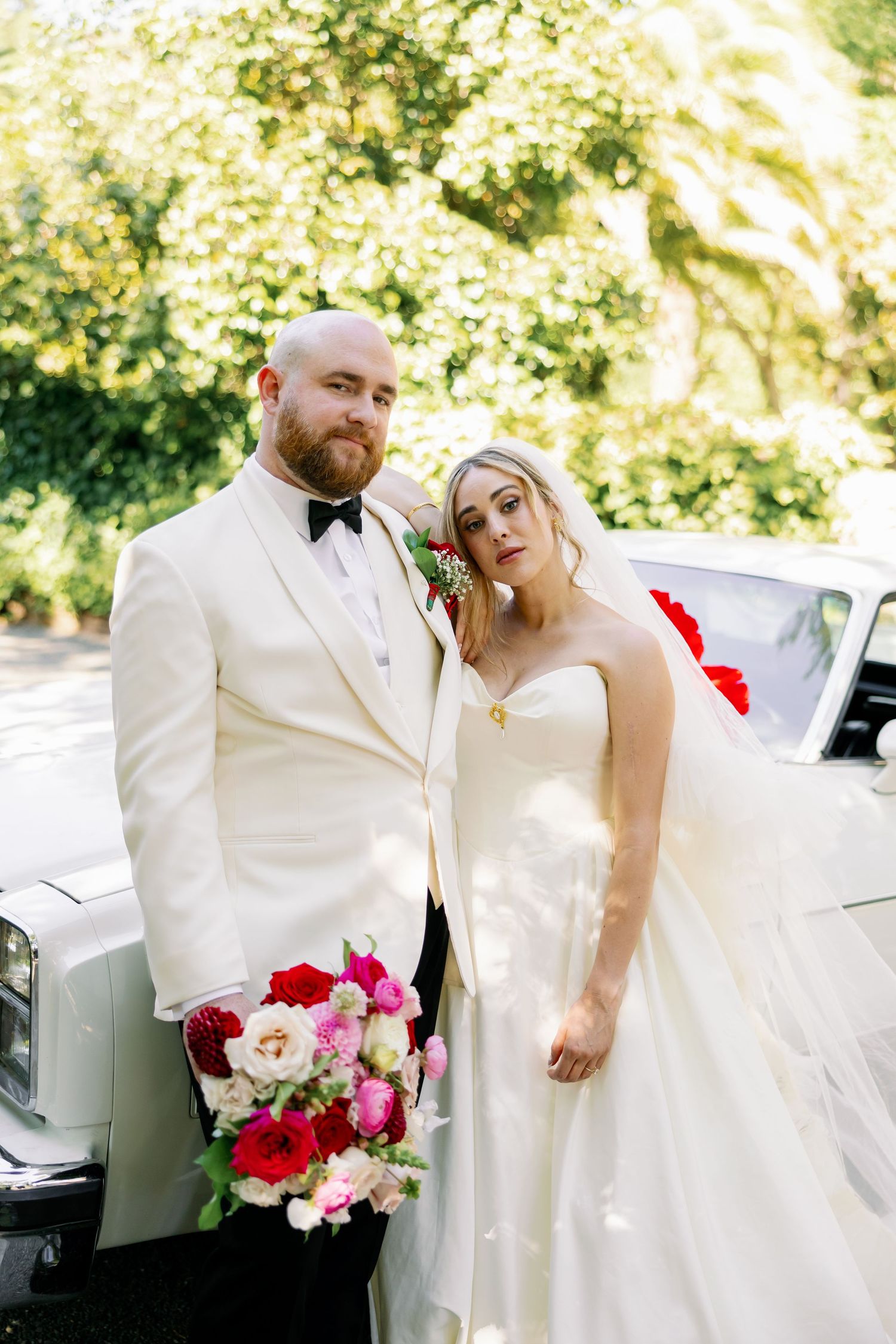 Bride and groom pose beside a white vintage car, she holds red and pink bouquet, he wears ivory tuxedo with bow tie.