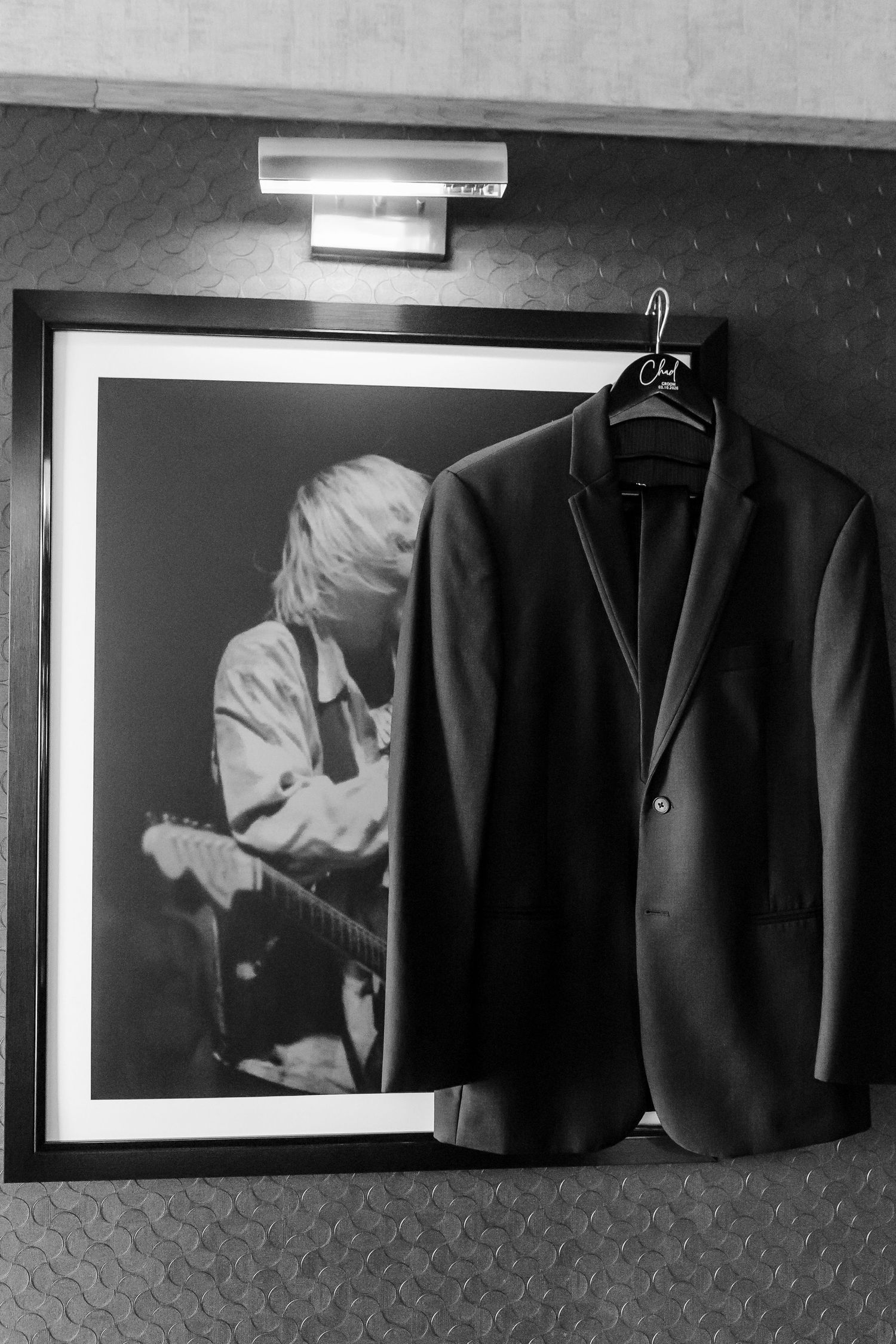 Black suit jacket on a hanger hanging over a framed black-and-white photo of a guitarist performing on stage, under a wall light.