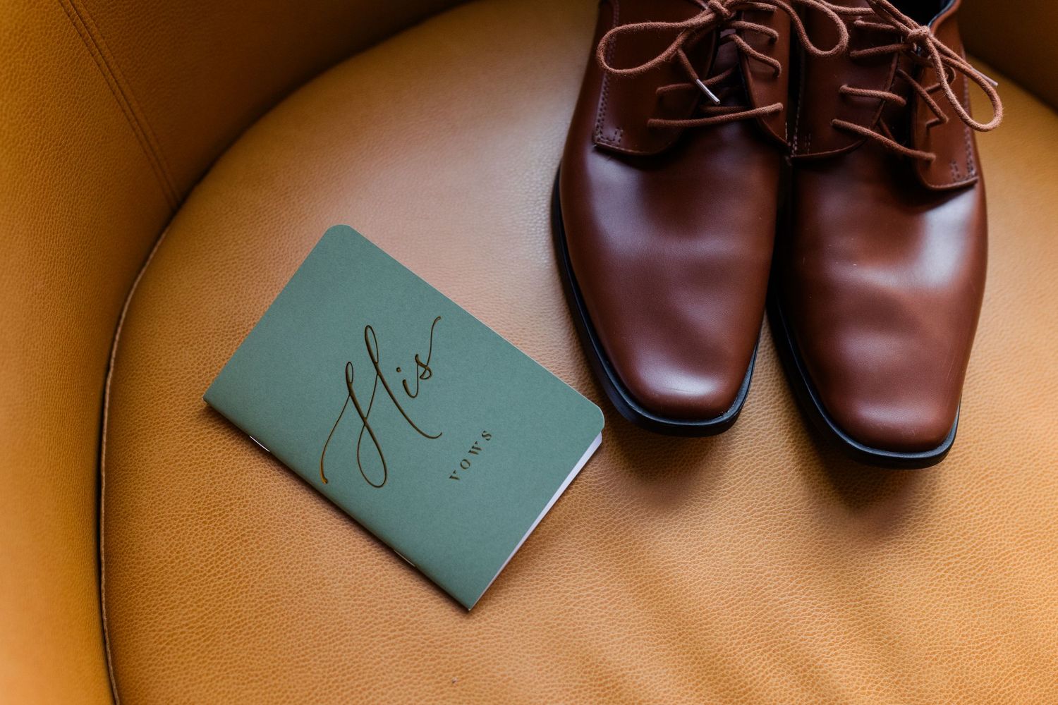 Polished brown leather dress shoes beside a green "His Vows" booklet resting on a tan leather chair, a groom's wedding day detail.