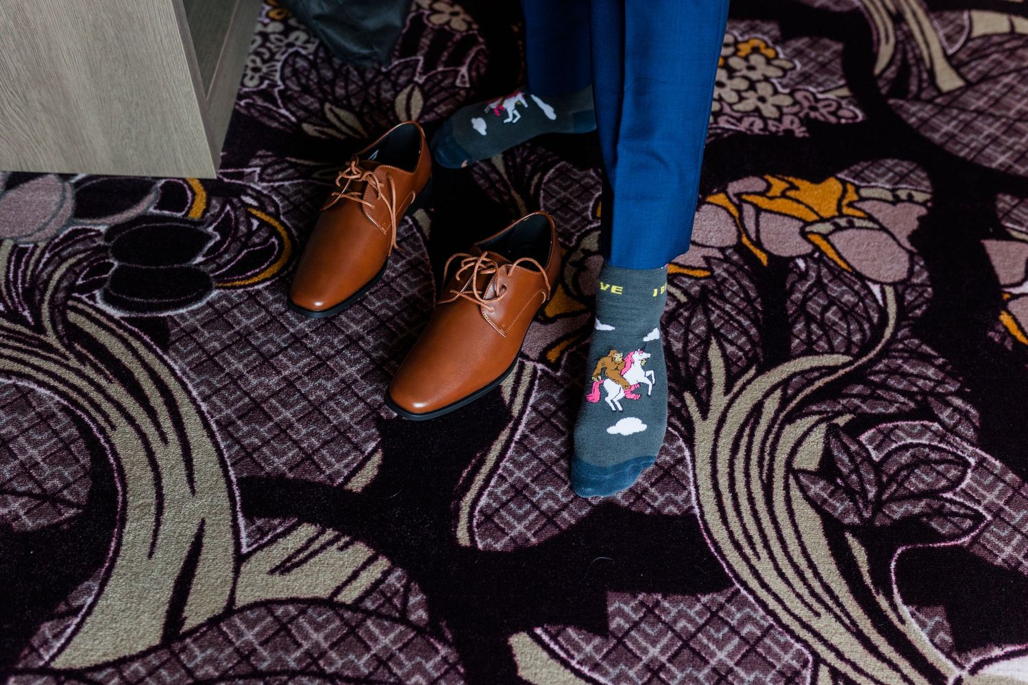 Person in blue pants and patterned socks stands beside a pair of brown leather dress shoes on a purple floral carpet.