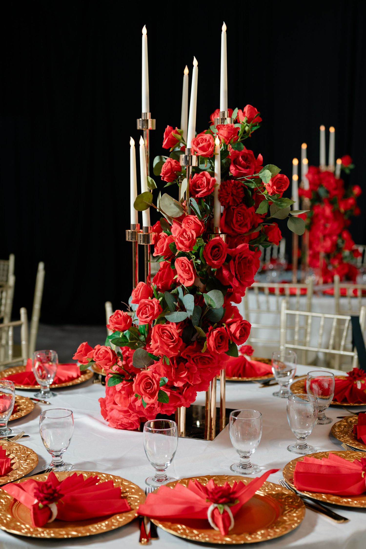 Dark blue castle-shaped cake on a white table surrounded by white candles with red ribbons, with a dessert table in the background.