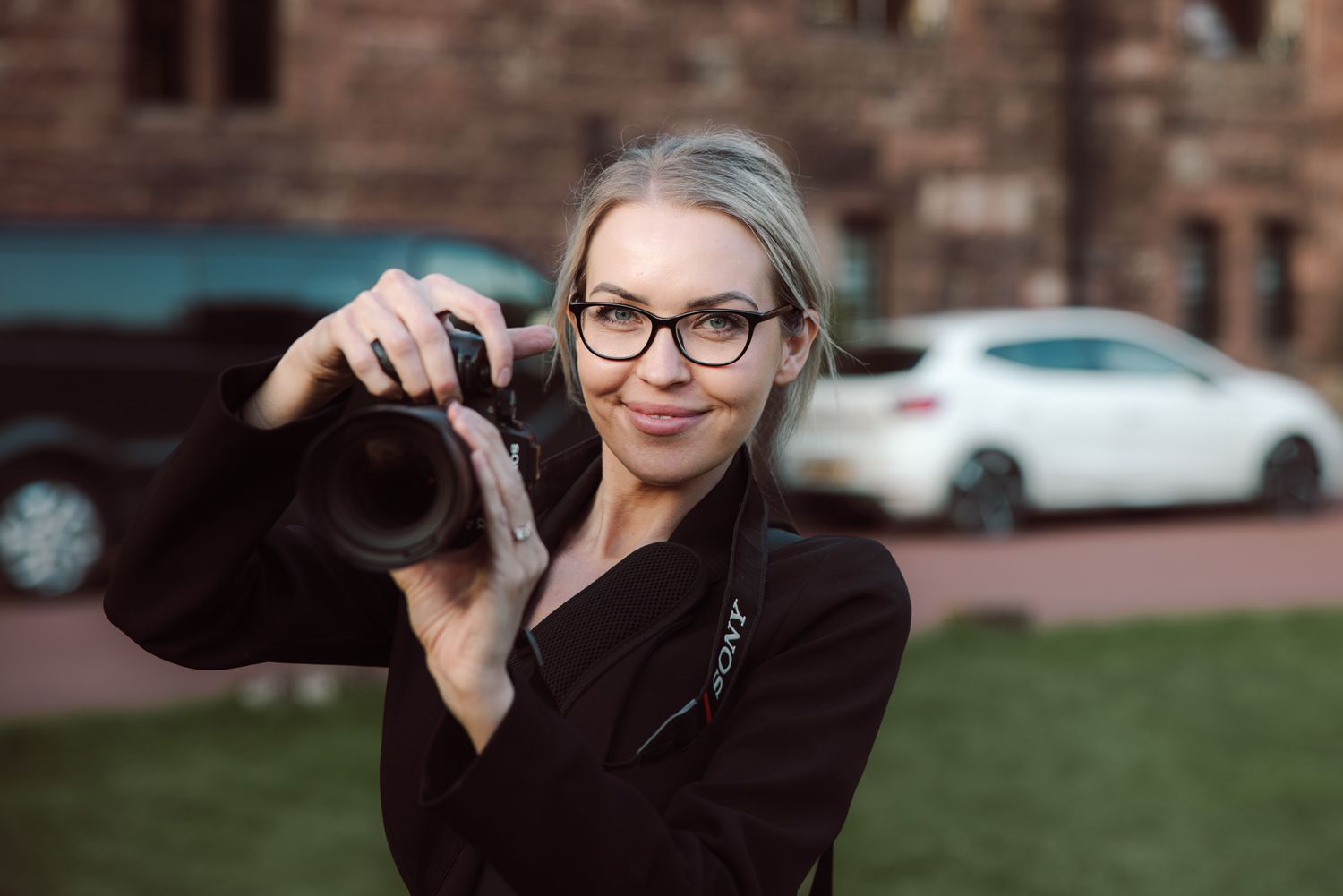Smiling woman with glasses and a black jacket holds a camera up to her eye outdoors near a brick building and parked cars.