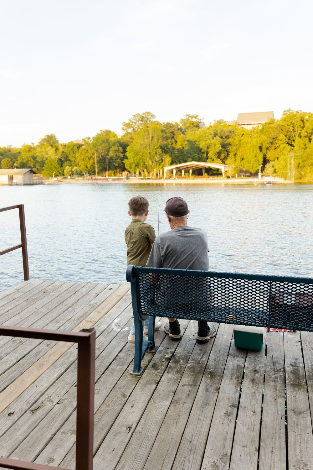 A man and a young boy sit on a bench on a wooden lakeside dock, fishing together at sunset with trees and a pavilion across the water.