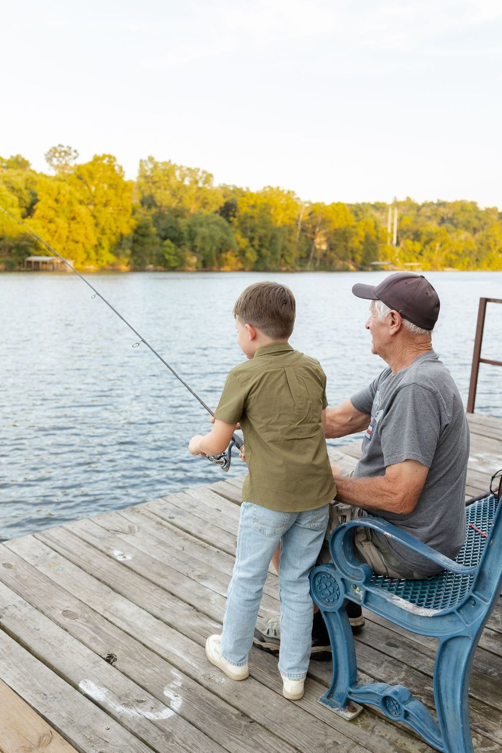 Grandfather and grandson fishing together from a wooden dock on a calm lake, with autumn trees lining the shore.