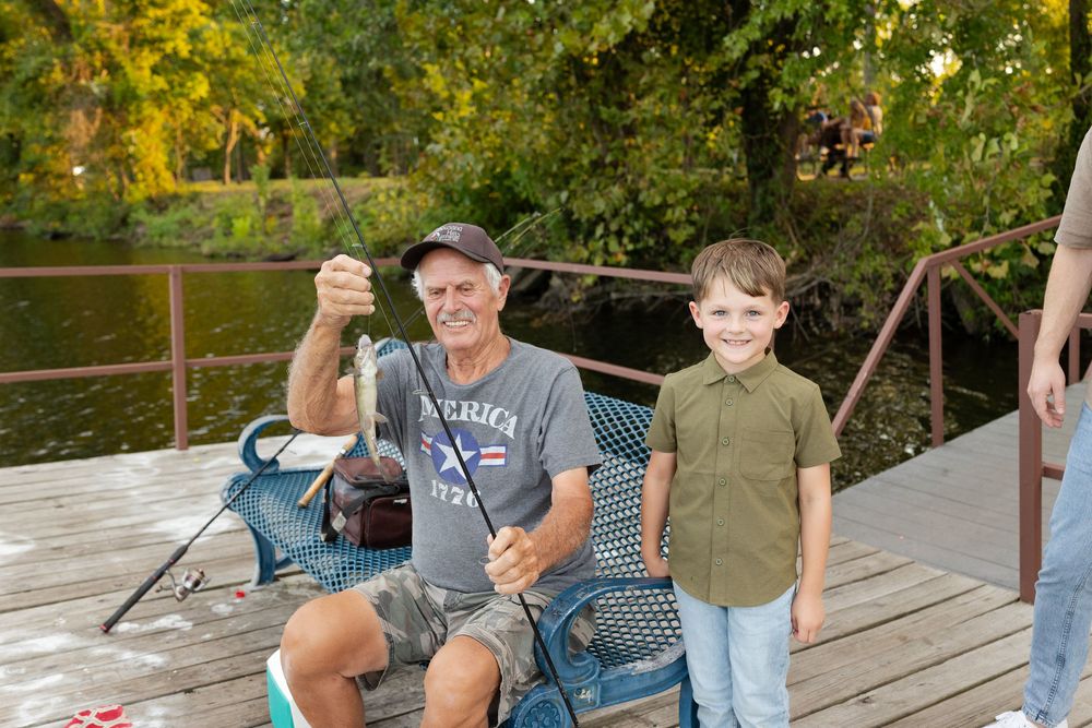 Smiling young boy stands beside a seated older man in a cap holding a fishing rod on a wooden dock by a calm lake.