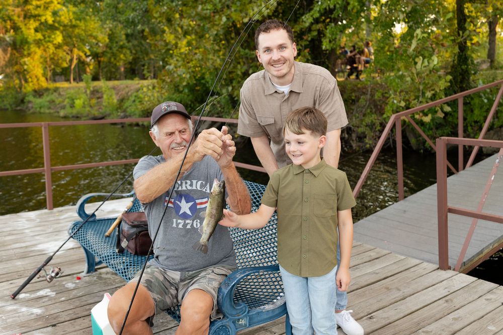 Three generations of a family smile on a wooden fishing dock as a grandfather holds up a freshly caught fish beside a boy and a man.