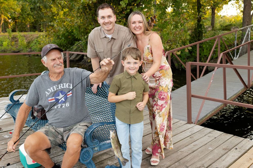 A smiling family of four poses on a wooden lakeside dock as a young boy holds a freshly caught fish beside an older man in a chair.