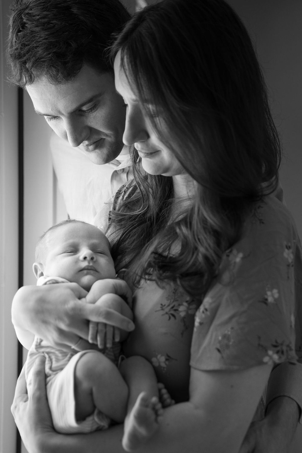 Black and white portrait of a mother and father gazing lovingly at their sleeping newborn baby while cradling the infant in their arms.