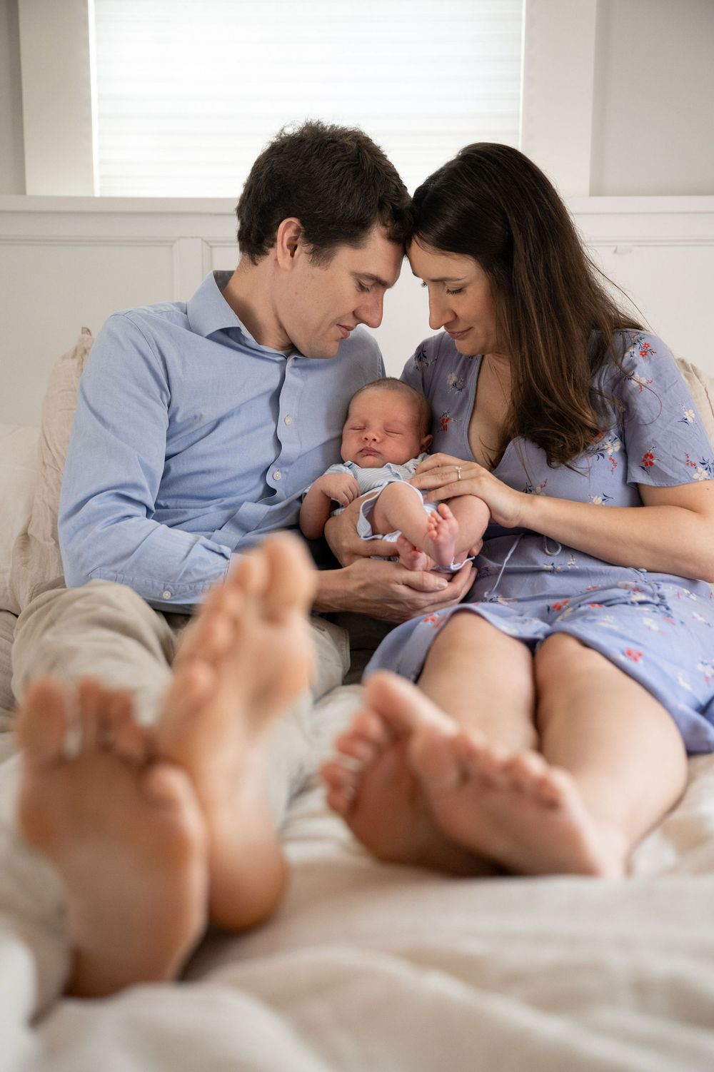 Smiling parents sit together on a bed gazing down at their sleeping newborn baby, with their bare feet in the foreground.