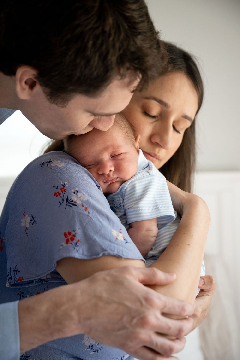 Parents tenderly embrace and gaze down at their sleeping newborn baby wrapped in a striped outfit, sharing a quiet, loving family moment.