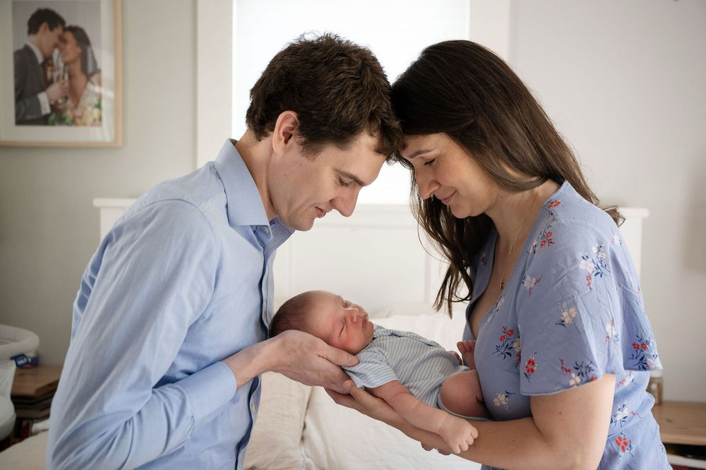 Smiling parents in light blue clothing gently cradle their sleeping newborn baby together in a bright, cozy room.