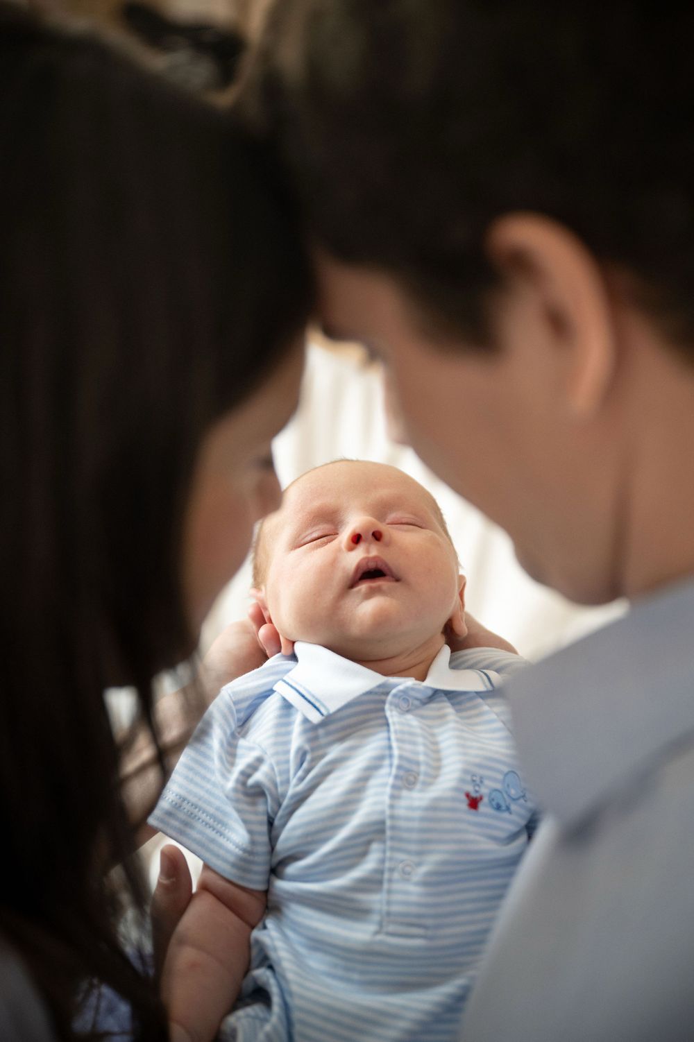 Two parents lean their foreheads together, gazing lovingly at a sleeping newborn baby in a blue striped polo shirt held between them.