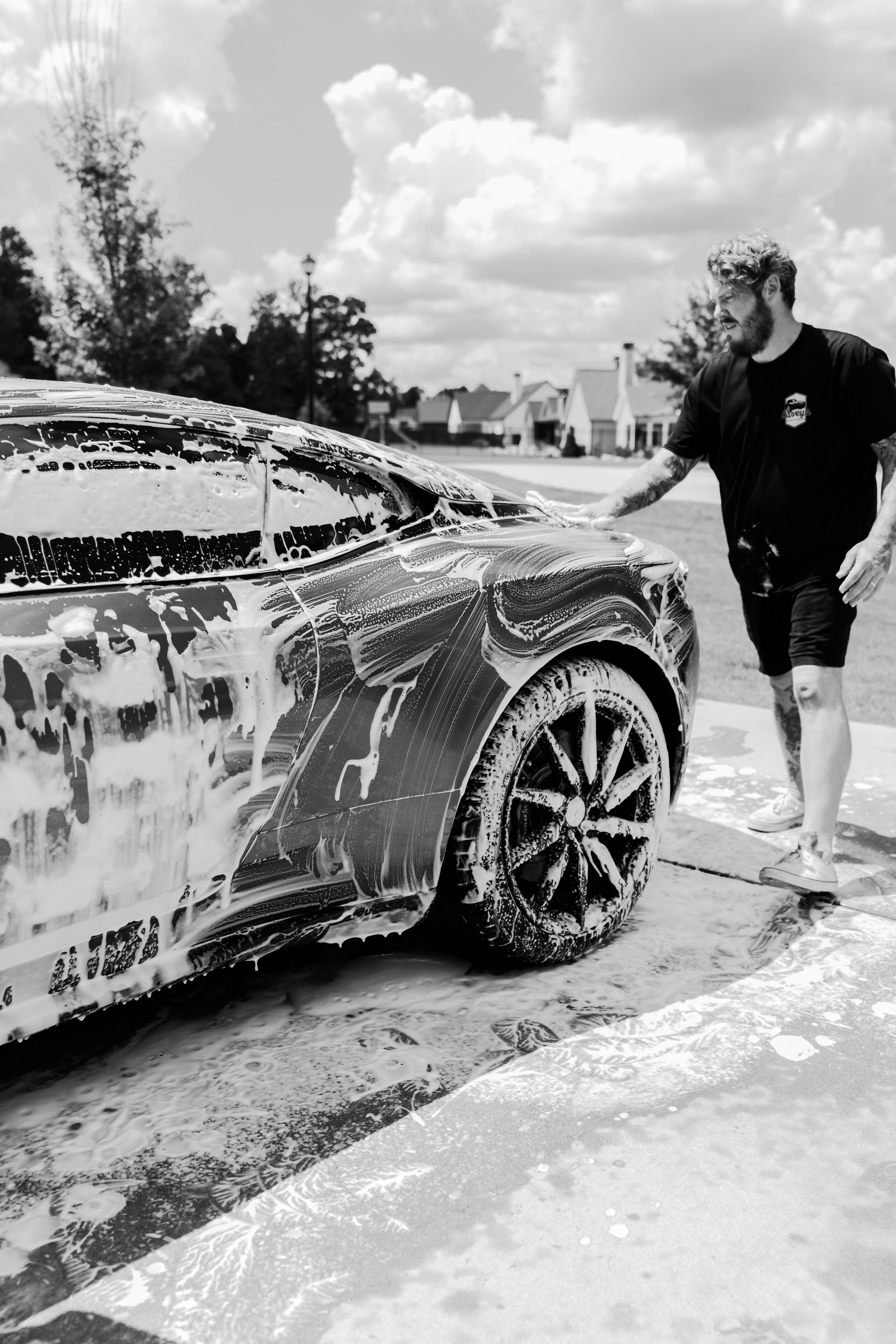 Man washing a luxury sports car covered in soap suds outdoors on a sunny day, black and white photo.