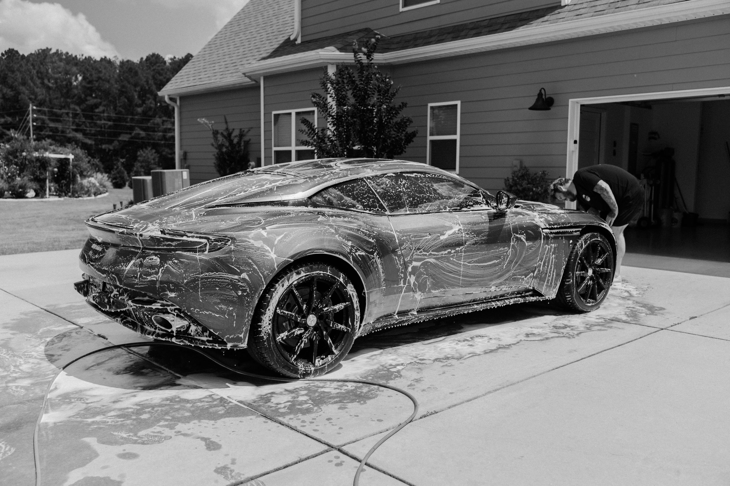 Black and white photo of a luxury sports car being hand-washed with soap suds in a residential driveway.