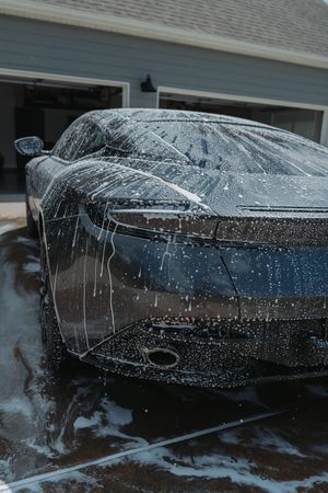 A sleek dark sports car covered in soap suds during a hand wash in a residential driveway.