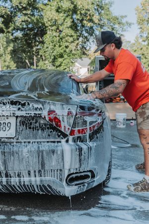A tattooed man in an orange shirt washes a soapy black sports car in a driveway on a sunny day.