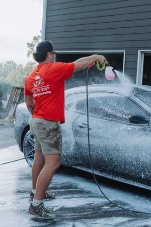 A car detailing professional in a red King shirt pressure washing a foam-covered black car outside a garage.